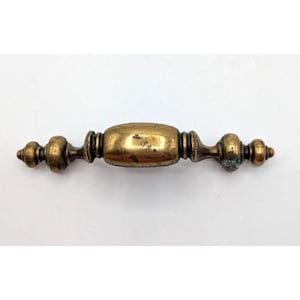 May include: Antique brass drawer pull with a central oval grip and decorative end details. The hardware has a tarnished, aged appearance, suggesting vintage or reclaimed use. The pull is designed for furniture restoration or adding a classic touch to cabinetry.