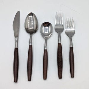 May include: A set of five pieces of silverware, including a knife, two spoons, and two forks. The utensils have stainless steel heads and dark brown wooden handles. The silverware is arranged on a white surface.