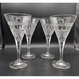 May include: Four clear crystal wine glasses with etched floral and geometric patterns. The glasses have a tall, slender stem and a wide, flared bowl. The glasses are arranged on a dark surface.
