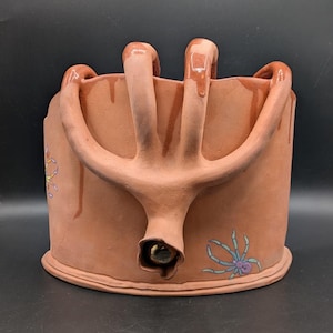May include: A handmade terracotta-colored ceramic bucket with a unique handle design. The bucket features a spout and is decorated with colorful spider illustrations. The handle has a dripped glaze effect.