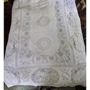 May include: A white rectangular tablecloth featuring an intricate lace pattern with floral motifs, central circular medallions, and decorative borders. The fabric displays a delicate openwork design throughout, creating a detailed texture.