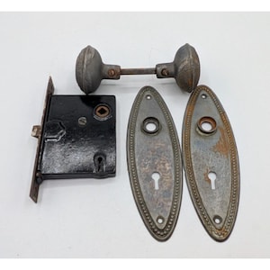 May include: An antique door hardware set featuring a black lock, two oval backplates, and a pair of doorknobs. The metal components have a weathered appearance, with signs of age and use. The backplates have a beaded edge design.