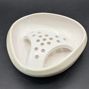 May include: A white ceramic flower frog with a curved, oval shape. The frog has a series of holes and is designed to hold flowers in place. The bowl is a creamy white color and has a smooth, glossy finish.