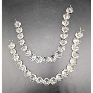 May include: Two strands of clear, faceted crystal beads. The beads are strung together with silver-colored wire. The strands are arranged in a curved shape, suggesting they could be used as a necklace or decorative element.