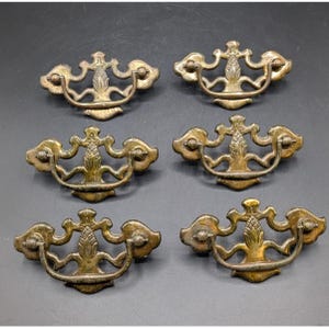 May include: A set of six brass-toned metal drawer pulls, each featuring a central pineapple motif and a curved, swinging handle. The hardware displays an aged, weathered finish with intricate scrollwork details on the backplate.