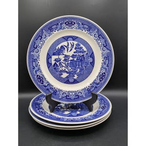 May include: A stack of three round ceramic plates featuring a blue and white Willow pattern. The design includes detailed illustrations of pagodas, trees, a bridge, and birds. The plates have a wide decorative border with geometric motifs.