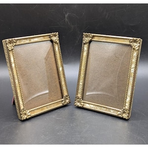 May include: Two antique gold-colored picture frames with ornate corner detailing. The frames have a rectangular shape with a light-colored interior, likely for displaying photos. The frames are set against a dark background.