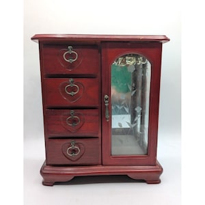 May include: A dark reddish-brown wooden jewelry armoire featuring four stacked drawers with heart-shaped metal pulls and a side cabinet with a glass door etched with a floral design. The cabinet includes a metal hook interior for hanging items.