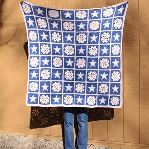 Puede incluir: Una manta de ganchillo azul y blanca con un patrón de estrellas y flores. La manta se sostiene frente a una pared de madera.