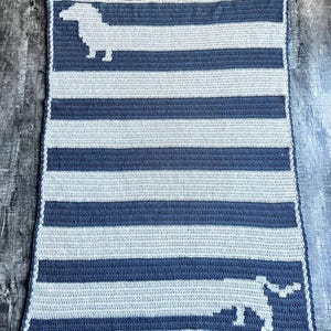 May include: A crocheted baby blanket with a striped pattern in shades of blue and white. The blanket features two dachshund silhouettes in white.