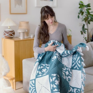 May include: A rectangular crochet blanket featuring a repeating geometric pattern in teal and white. The blanket is constructed from multiple square panels with intricate stitch work and is finished with white fringe along the side edge.