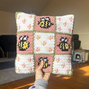 May include: A square crochet pillow featuring a grid pattern of nine alternating squares. The design includes white squares with small pink and yellow flower motifs and dusty pink squares displaying black and yellow bumblebee graphics, all bordered by olive green yarn.