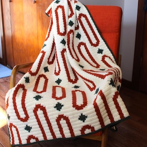 May include: A cream-colored crocheted blanket with a repeating pattern of rust-colored elongated ovals and dark green diamond shapes. The blanket is draped over an orange chair with a wooden frame. The edges are finished with a dark green border.