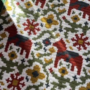 May include: Hand-crocheted blanket with a repeating pattern of red Dala horses, yellow flowers, and red starbursts on a white background. Green and blue accents are also present. The blanket has a textured appearance.