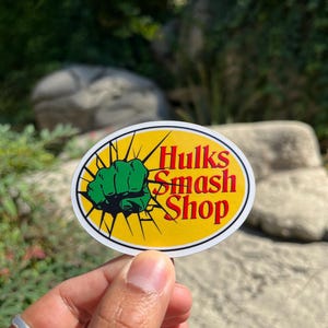 Può includere: Adesivo ovale con sfondo giallo e testo "Hulks Smash Shop" in rosso e bianco. Un pugno verde è a sinistra. L'adesivo è tenuto davanti a uno sfondo sfocato verde e grigio.