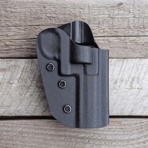 Könnte beinhalten: Ein schwarzes, geformtes Polymerholster mit drei sichtbaren Schrauben. Das Holster hat eine strukturierte Oberfläche und ist für die sichere Aufbewahrung einer Schusswaffe konzipiert. Das Design umfasst eine Gürtelschlaufe und eine konturierte Form.