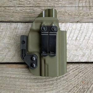 Könnte beinhalten: Eine olivgrüne Holstertasche mit schwarzen Metallclips und Beschlägen. Die Holstertasche besteht aus einem strapazierfähigen Material und hat eine strukturierte Oberfläche. Das Design umfasst einen Gürtelclip zur sicheren Befestigung.