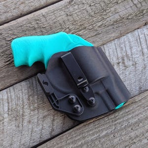 Könnte beinhalten: Eine schwarze Polymerholster mit einem schwarzen Metallclip und Schrauben. Ein türkisfarbener Pistolenhandgriff ist im Inneren der Holster sichtbar. Die Holster liegt auf einer verwitterten Holzoberfläche.