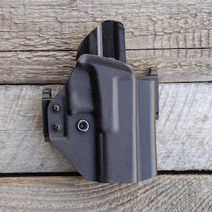 Könnte beinhalten: Schwarze Polymerholster mit strukturierter Oberfläche. Das Holster verfügt über einen Gürtelclip und wird mit Schrauben und einer Metallöse befestigt. Das Design ist so geformt, dass es zu einer Schusswaffe passt und sicheres Tragen ermöglicht.