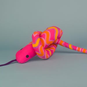 May include: A colourful plush snake toy with a pink head, black eyes, and a purple tongue. The body is patterned with orange and yellow stripes. The background is a light blue.