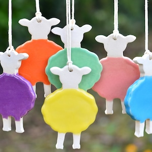 Könnte beinhalten: Sechs Keramik-Schaf-Anhänger, die an weißen Schnüren hängen. Die Schafe sind weiß mit unterschiedlich gefärbten Körpern: lila, orange, grün, rosa, gelb und blau.