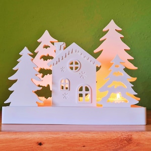 Puede incluir: Una escena de pueblo navideño decorativa blanca, con una casa y varios abetos estilizados. La casa tiene ventanas y una chimenea. La escena está iluminada desde dentro, creando un brillo cálido. El fondo es verde liso.