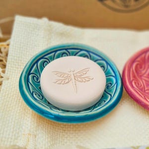 May include: A round, pale soap with a dragonfly design sits in a teal ceramic dish. The dish features a decorative, swirling pattern around the edge. The soap dish is placed on a cream-coloured cloth.