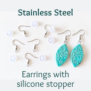 Op de afbeelding: Een paar turquoise bladvormige oorbellen met een wit bloemmotief, vergezeld van vier roestvrijstalen oorhaken en vier doorzichtige siliconen stoppers. De tekst Stainless Steel Earrings with silicone stopper is zichtbaar.