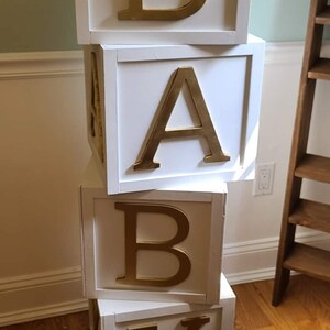 May include: A stack of four white wooden blocks with gold letters spelling out the word "BABY".