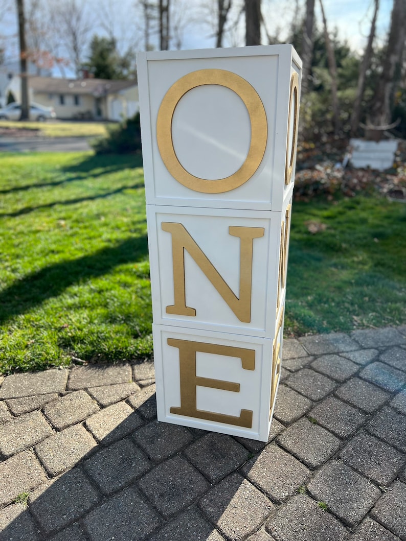 May include: White wooden blocks stacked to spell "ONE" with gold letters. The blocks are on a gray brick surface.