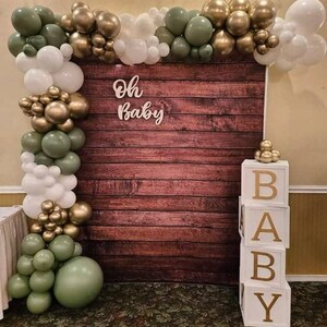 May include: A wooden backdrop with a green, gold, and white balloon garland. The backdrop has the words "Oh Baby" written on it. There are also white blocks that spell out "BABY" in gold letters.