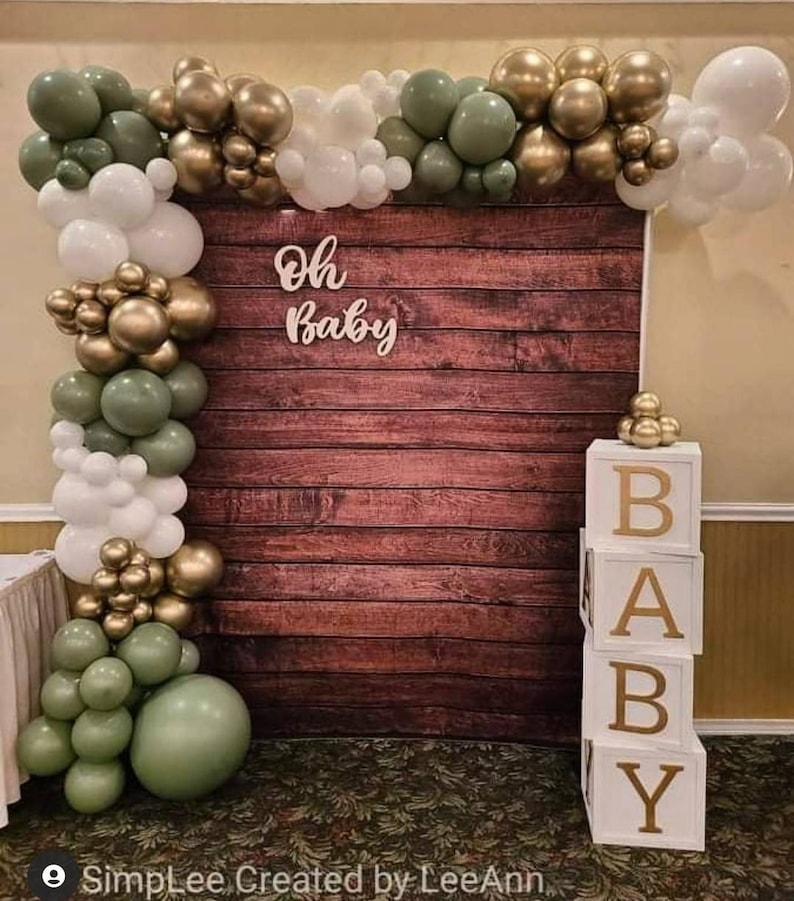 May include: A wooden backdrop with a green, gold, and white balloon garland. The backdrop has the words "Oh Baby" written on it. There are also white blocks that spell out "BABY" in gold letters.