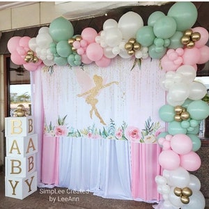 May include: A baby shower backdrop with a fairy silhouette, pink and white floral design, and a white and pink curtain. The backdrop is decorated with a garland of green, white, and pink balloons.