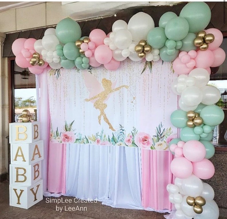 May include: A baby shower backdrop with a fairy silhouette, pink and white floral design, and a white and pink curtain. The backdrop is decorated with a garland of green, white, and pink balloons.