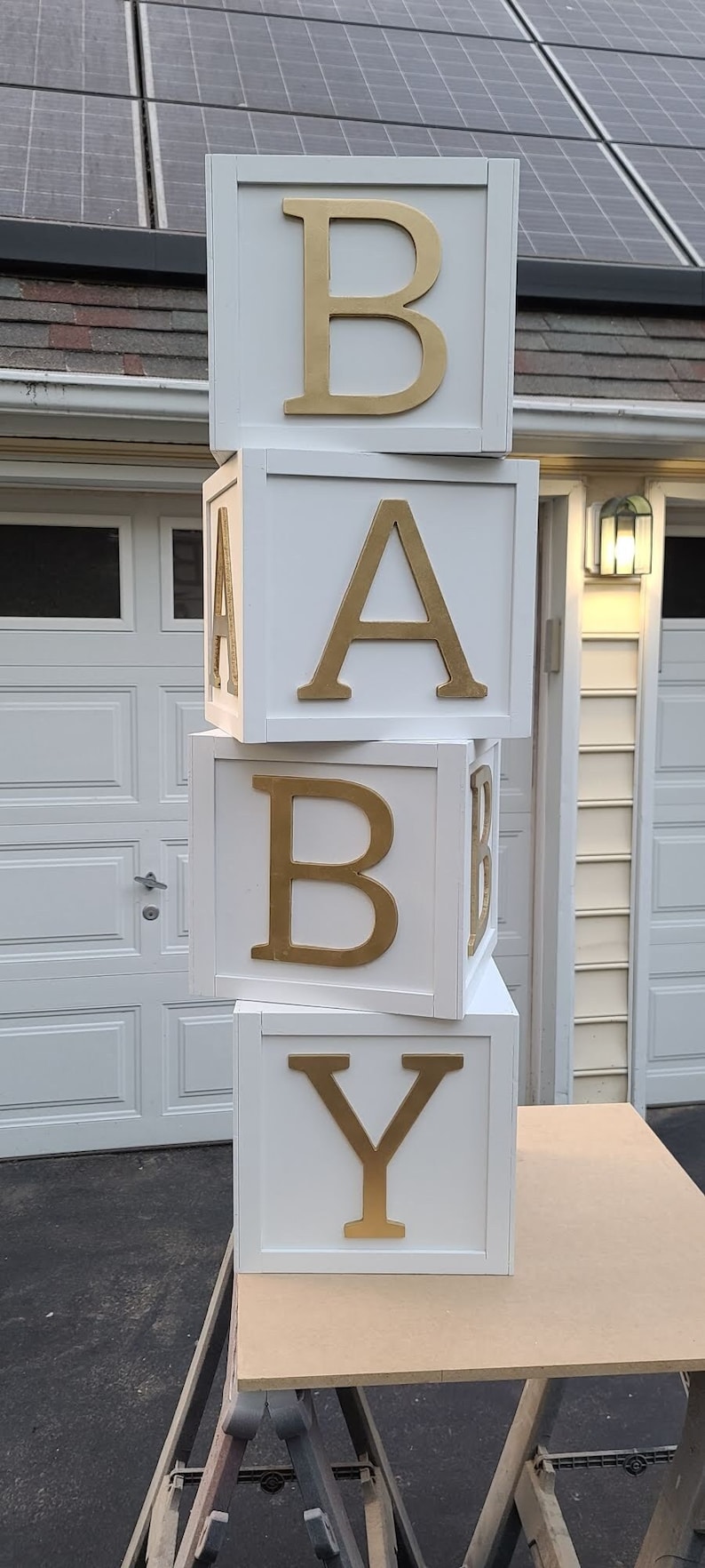May include: Four white wooden blocks stacked on top of each other, spelling out the word "BABY" in gold letters. The blocks are sitting on a wooden table.