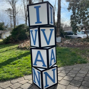 May include: A stack of four white blocks with black borders and blue letters spelling out the name "IVAN". The blocks are sitting on a brick patio.