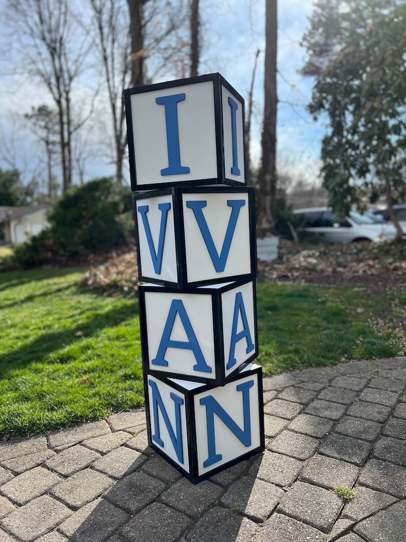 May include: A stack of four white blocks with black borders and blue letters spelling out the name "IVAN". The blocks are sitting on a brick patio.