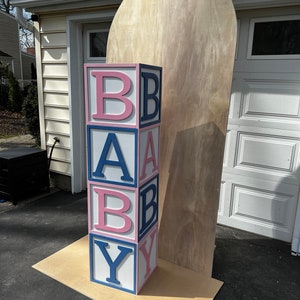 May include: A stack of large, colorful blocks spelling out "BABY" in pink and blue letters. The blocks are stacked on a wooden platform in front of a wooden backdrop.