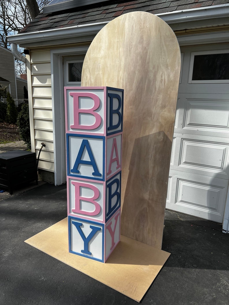 May include: A stack of large, colorful blocks spelling out "BABY" in pink and blue letters. The blocks are stacked on a wooden platform in front of a wooden backdrop.