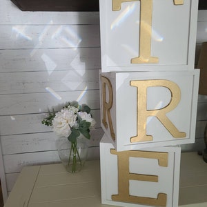 May include: Three white wooden blocks stacked on top of each other. Each block has a gold letter on the front. The letters spell out the word "TRE".