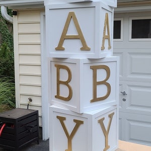 May include: White wooden blocks stacked to spell "BABY" with gold letters. The blocks are sitting on a wooden table.