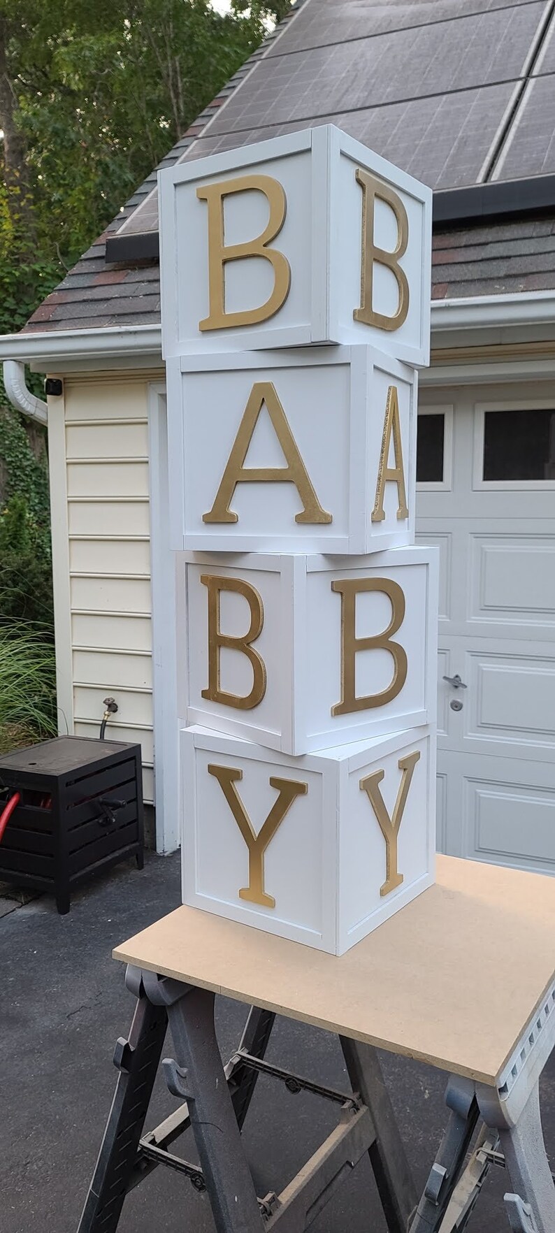 May include: White wooden blocks stacked to spell "BABY" with gold letters. The blocks are sitting on a wooden table.