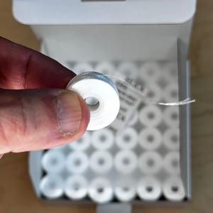 Peut inclure: Une boîte de bobines de fil dentaire blanches. Les bobines sont disposées en grille et sont toutes de la même taille. Une bobine est tenue par une main.