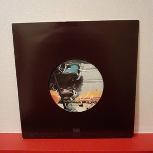 Może przedstawiać: Czarny 7-calowy winylowy singiel z kolorową etykietą przedstawiającą samochód jadący ulicą. Etykieta zawiera napis "OMD" i "Souvenir".