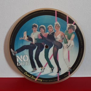 May include: A 7-inch vinyl record featuring a band called "No Dice". The band members are dressed in colourful clothing and are striking a dynamic pose. The record is titled "Come Dancing" and was released in 1979.