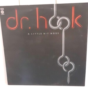 Puede incluir: Una portada de álbum de vinilo negro con el título "dr. hook" en letras rojas de neón y las palabras "A LITTLE BIT MORE" en letras blancas debajo. La portada del álbum presenta un diseño de gancho plateado.