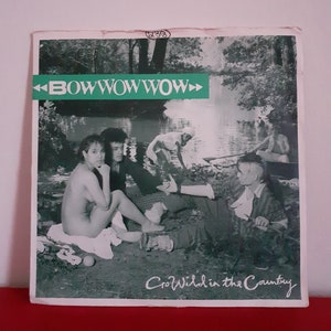 May include: A black and white photo of a band called "Bow Wow Wow" on a record album cover. The band members are sitting in a forest by a lake. The album title is "Go Wild in the Country".