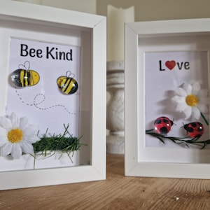 Könnte beinhalten: Zwei gerahmte Kunstwerke in weißen Rahmen. Eines zeigt "Bee Kind" mit Bienen- und Gänseblümchen-Akzenten. Das andere zeigt "Love" mit Marienkäfer- und Gänseblümchen-Akzenten. Beide haben einen weißen Hintergrund.
