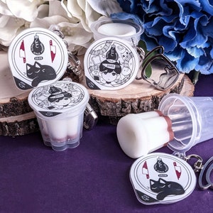 May include: Four black and white illustrated keychains with different designs. The keychains are attached to clear plastic containers. The containers are on a wooden surface with a purple background.