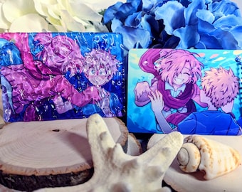 May include: Two anime-style character charms featuring a pink-haired character with a blue scarf and a blonde-haired character. The charms are on a wooden surface with a seashell and a starfish.